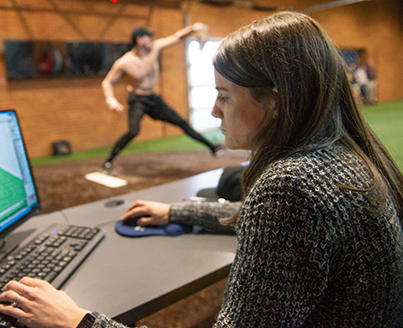 Female researcher studies data on laptop while baseball player pitches behind her in new pitching lab