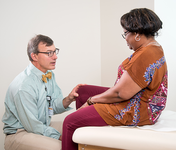 white male wearing bow tie examines knee of African American female patient sitting on exam room bed