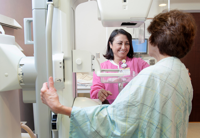 A smiling Latina woman in a pink top positions equipment for a mammogram on a brunette woman wearing a hospital gown