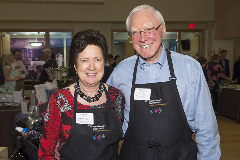 An older man with white hair and woman with brown hair, both wearing black aprons with the Brenner Children's logo, smile at the camera