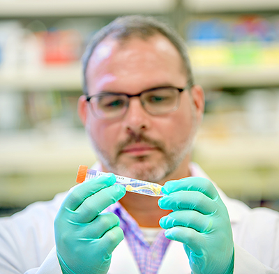 A man in glasses, gloves and a white coat looks at a test tube he is holding